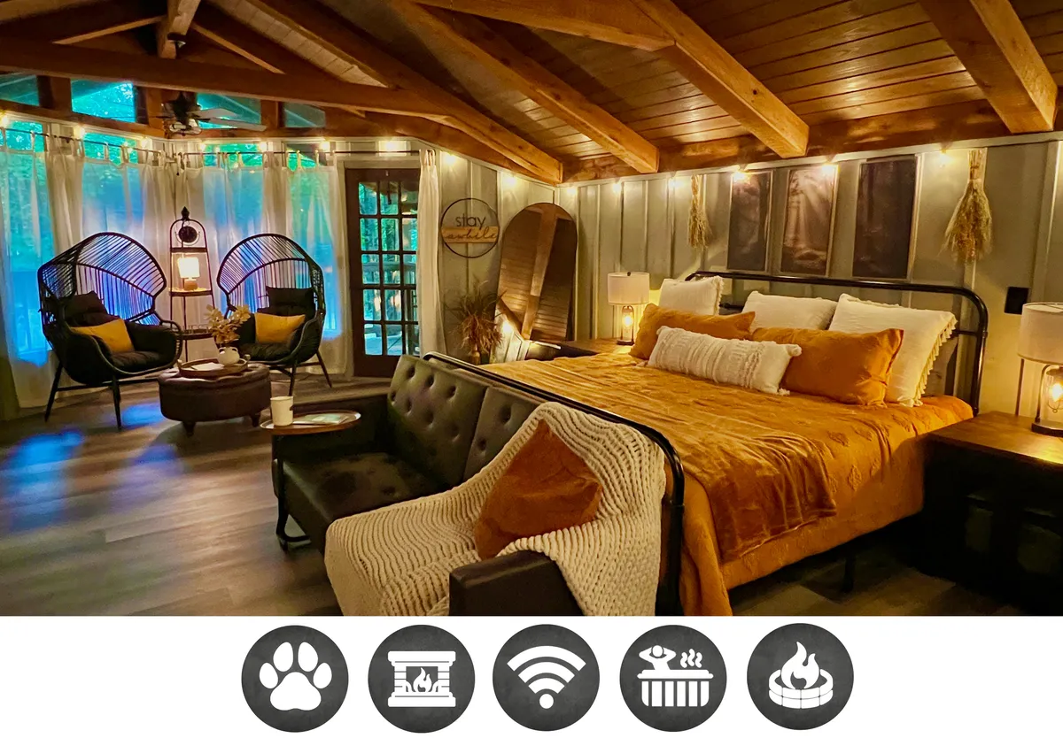 The image showcases the inviting interior of the Light'n Bug Bungalow, featuring a plush, cozy bed, unique hanging chairs perfect for unwinding, rustic wooden beams overhead, and warm, ambient lighting. Large windows offer views for stargazing, while a fireplace and soft furnishings create an atmosphere ideal for relaxing evenings, reading, or intimate conversations.