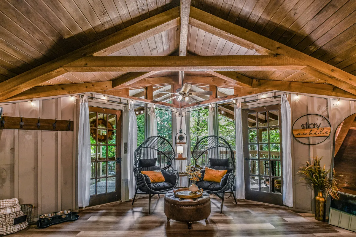Here, two large hanging chairs and a round coffee table are positioned by expansive windows with curtains, featuring a sign that reads 'stay awhile.' The tranquil setting captures the essence of unwinding in a cabin surrounded by nature, with soft natural light filtering in and comfortable seating that invites guests (and their pets) to relax after a day outside.