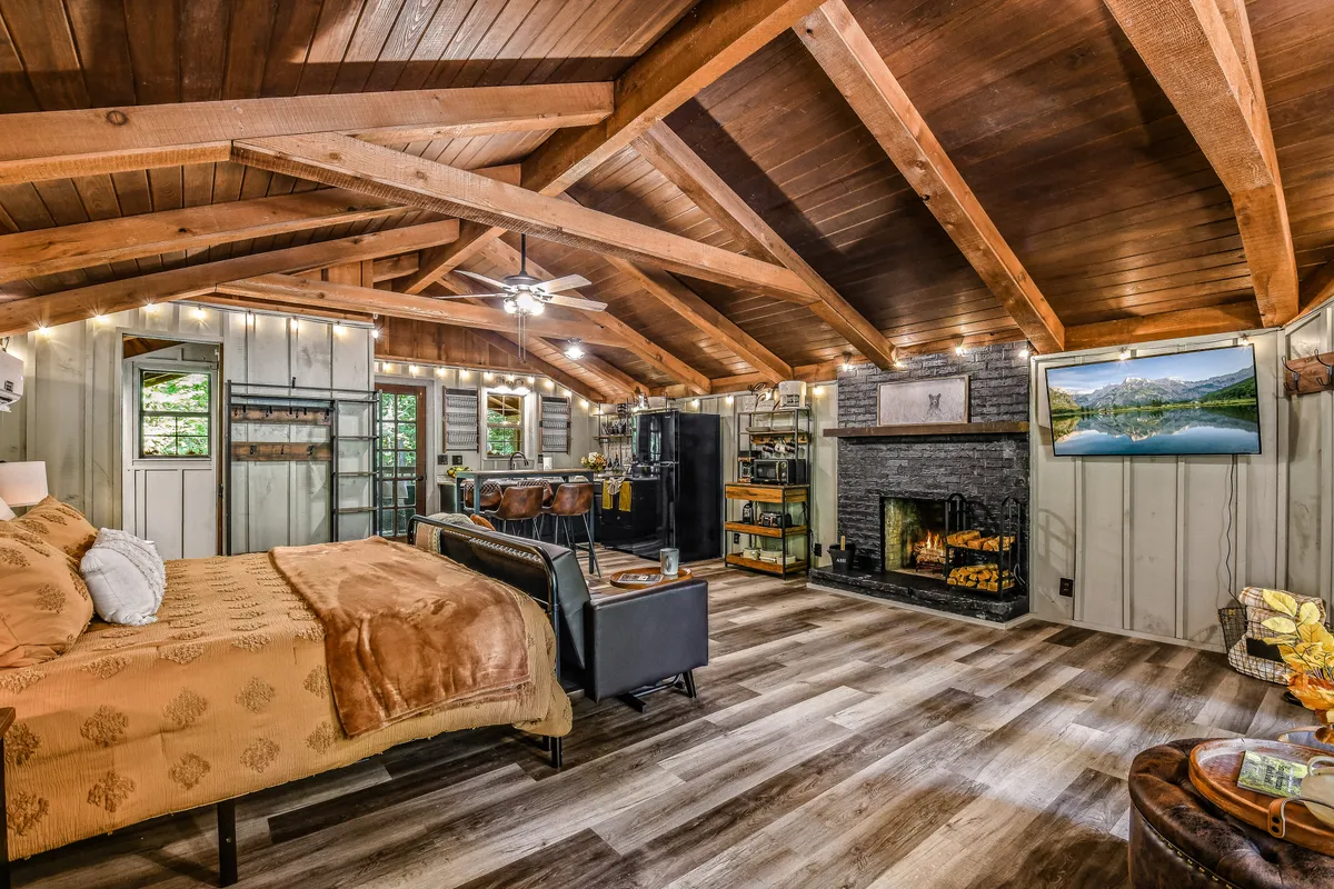 This image captures a warm, open studio layout featuring a king bed with decorative pillows, a cozy fireplace, a flat-screen TV positioned for easy viewing, and a dining area for four. The wooden beams and soft lighting create a welcoming, unified atmosphere, highlighting the seamless flow between the sleeping, dining, and living spaces.