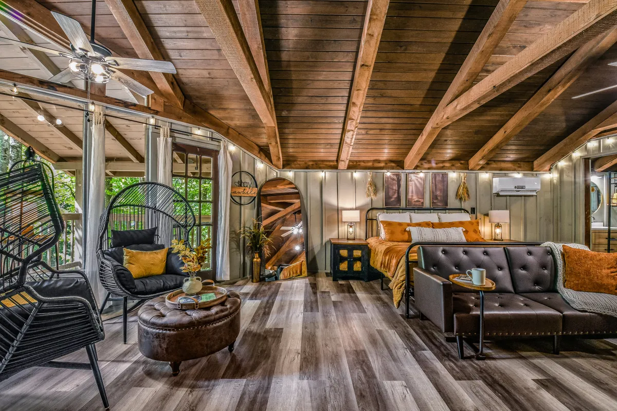 This image features a cozy bed with decorative throw pillows, stylish armchairs set up for conversation or relaxation, a large full-length mirror, wooden beams overhead, and plenty of natural light. The arrangement encourages both intimate moments and shared experiences, perfectly matching the studio’s flexible sleeping and living setup.