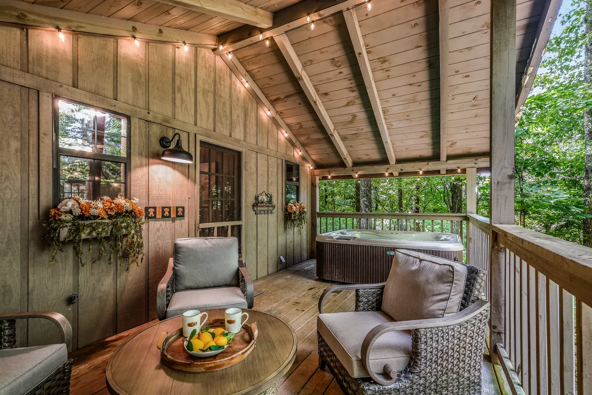 A welcoming exterior porch space with a hot tub, ample seating, decorative plants, and warm lighting fixtures. The area is designed for both relaxation and socializing, with views of the surrounding woods.
