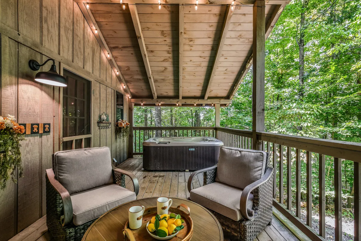A tranquil exterior deck featuring a private hot tub, a cozy seating area with a table adorned with fruit, and soft decorative lighting. The space is enveloped by lush trees, offering an intimate and secluded atmosphere ideal for relaxing and unwinding after a day exploring the mountains.