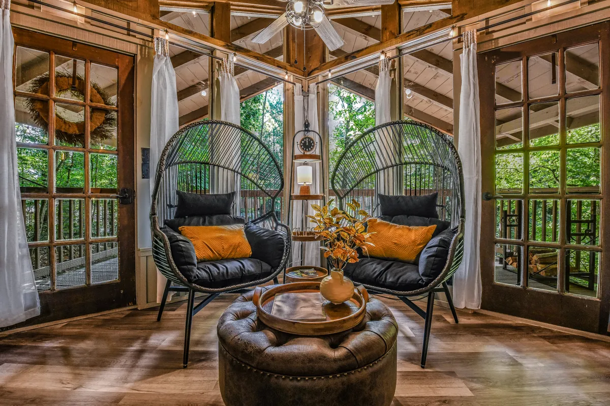 This image shows a comfortable living area with large windows that let in morning light, hanging chairs, a coffee table, and decorative plants. The view outside the windows reveals lush, seasonal foliage. The space feels open and connected to the outdoors, suggesting a seamless transition between indoor relaxation and outdoor adventure.