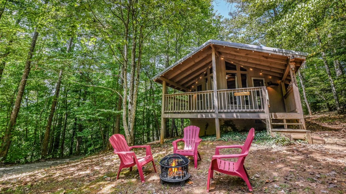 An exterior scene featuring a charming wooden cabin, a fire pit surrounded by red chairs, and a forested background. The setup is ideal for campfire gatherings, stargazing, and sharing stories under the night sky.