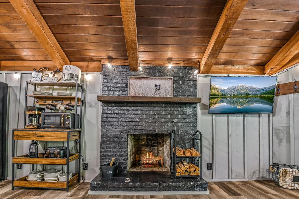 This image features a cozy fireplace with stacked wood, a TV that offers a view out towards the mountains, and a shelf with kitchen items. The room feels warm, inviting, and connected to the surrounding nature, especially through the prominent window view.
