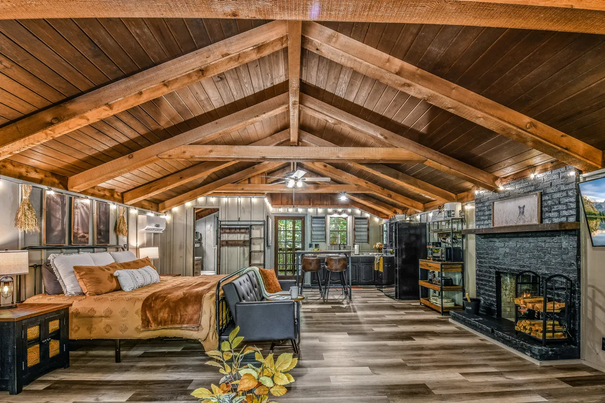 A warmly lit interior features a cozy fireplace, wooden beams, a comfortable bed with decorative pillows, and a bar area perfect for gatherings. The inviting space is ideal for relaxing evenings, family bonding, or enjoying a peaceful morning together.